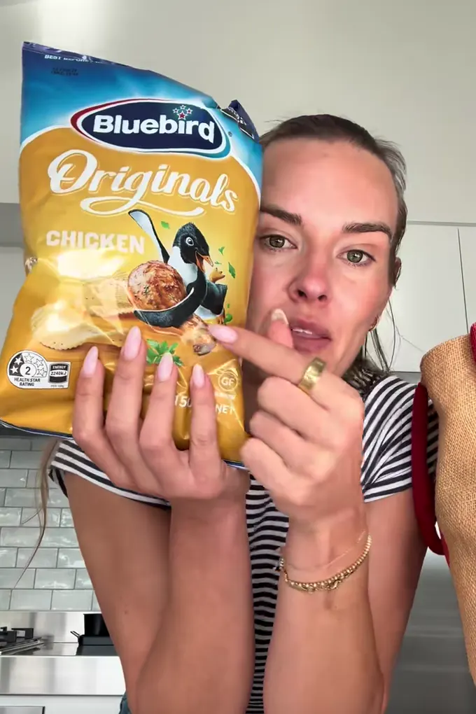 Example post, a photo: a woman leaning on her kitchen bench holding up a bag of chicken chips